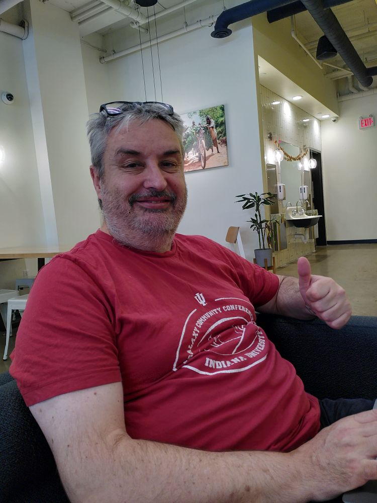 Simon Gladman is sitting on a couch, he looks absolutely happy as can be, staring into the camera and giving a thumbs up. He has salt and pepper hair and a scruffy grey beard that look like he hasn't slept in a few days. He is wearing his firetruck red indiana galaxy community conference shirt.